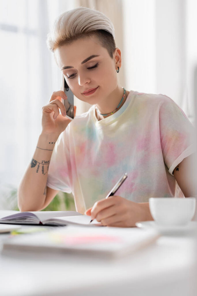 stylish woman writing in notebook and talking on mobile phone near blurred coffee cup - Фото, изображение