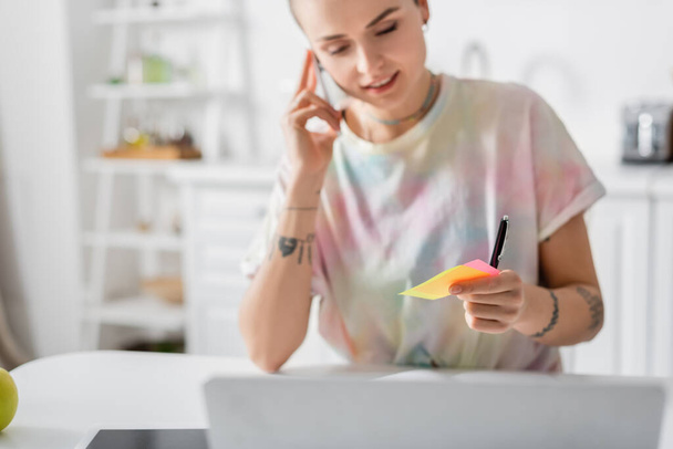 positive woman holding sticky notes while talking on smartphone near laptop - Foto, Imagem
