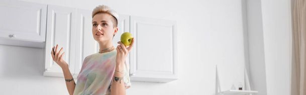 smiling woman with trendy hairstyle holding apple and pen in kitchen, banner - Photo, image