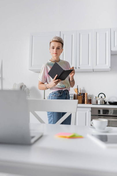stylish woman standing with notebook in kitchen and looking at blurred laptop - Fotó, kép