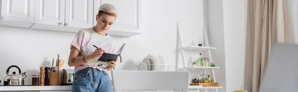 young woman with trendy hairstyle writing in notebook in kitchen on blurred foreground, banner - Foto, Imagen