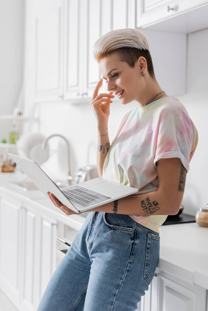 cheerful woman gesturing while standing with laptop in kitchen - Fotoğraf, Görsel