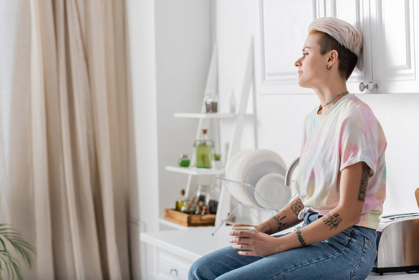 young tattooed woman sitting on worktop in kitchen and looking away - Foto, Bild
