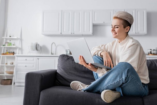 happy woman looking at laptop while sitting on couch with crossed legs - Фото, изображение
