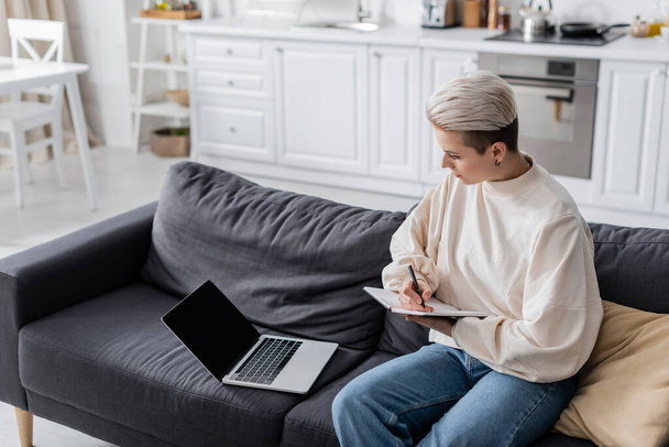 woman with trendy hairstyle writing in notebook near laptop on sofa at home - Zdjęcie, obraz