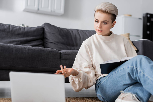 woman in white pullover sitting on floor with notebook and pointing at blurred laptop - Φωτογραφία, εικόνα