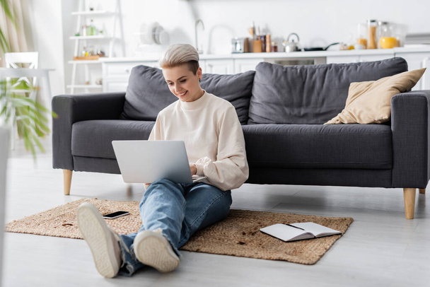 longitud completa de la mujer con el ordenador portátil sentado en el suelo cerca del sofá, portátil y teléfono móvil - Foto, imagen