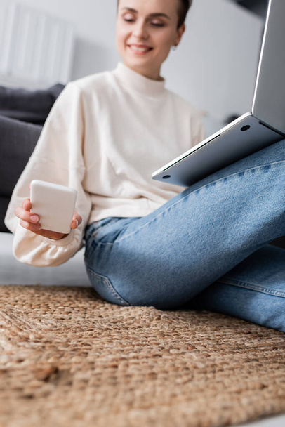 blurred woman looking at smartphone while sitting on floor with laptop - Foto, Imagem