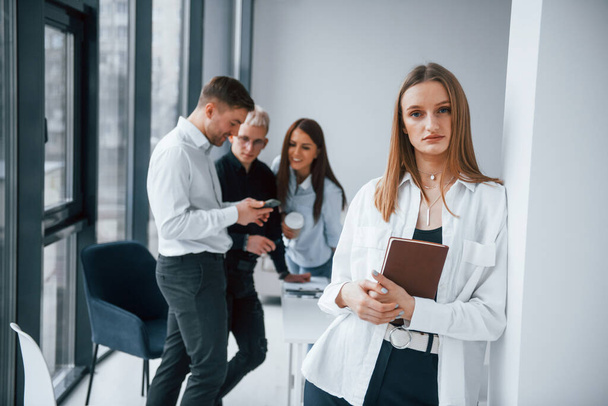 Portrait of cheerful blonde with notepad that stands in front of young successful team that working and communicating together indoors in office. - 写真・画像