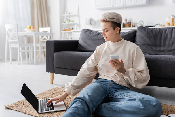 positive woman with mobile phone sitting on floor and using laptop with blank screen - Fotoğraf, Görsel
