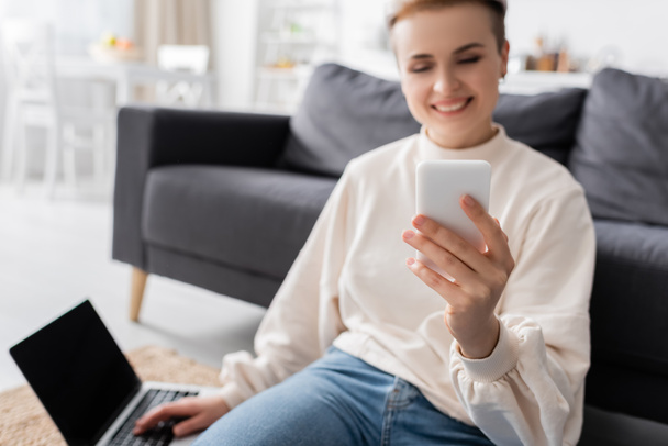 cheerful woman sitting on floor near laptop and looking at mobile phone, blurred background - Foto, imagen