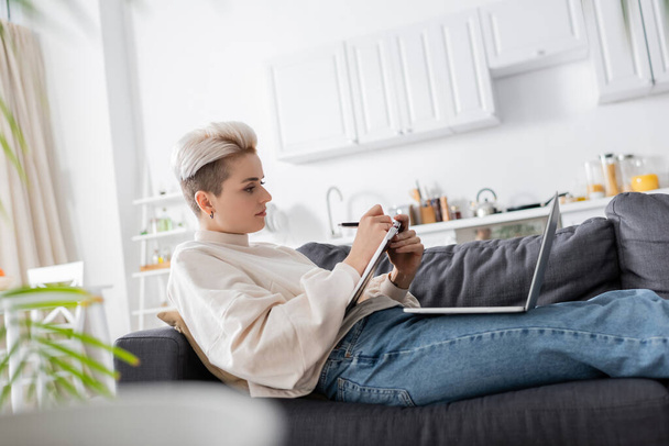 side view of woman writing in notebook near laptop on sofa at home - Photo, Image