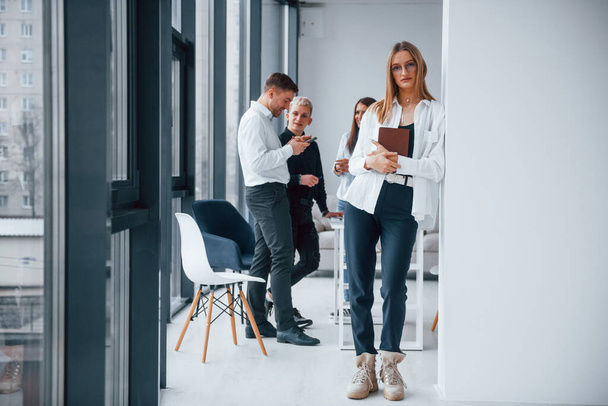 Portrait of cheerful blonde with notepad that stands in front of young successful team that working and communicating together indoors in office. - Фото, зображення
