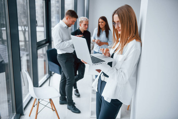 Blonde with laptop stands in front of young successful team that working and communicating together indoors in office. - Фото, зображення