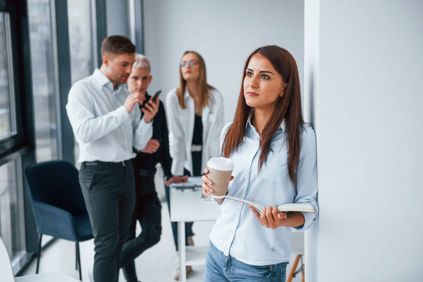 Portrait of cheerful woman with cup of drink that stands in front of young successful team that working and communicating together indoors in office. - Φωτογραφία, εικόνα