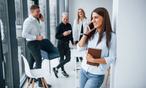 Portrait of cheerful woman with cup of drink and phone that stands in front of young successful team that working and communicating together indoors in office. - Photo, image