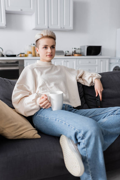 trendy woman in white pullover holding cup of tea and watching movie on tv at home - Foto, Imagem