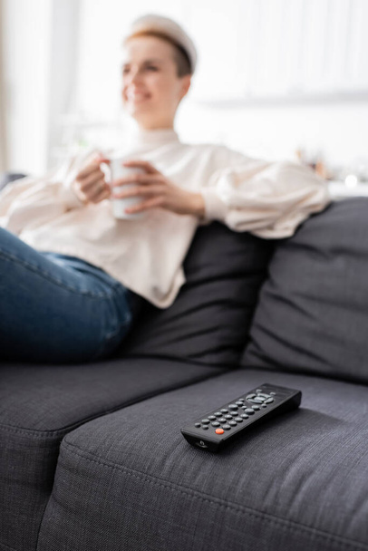 partial view of blurred woman with cup of tea sitting on couch near tv remote controller - Fotografie, Obrázek