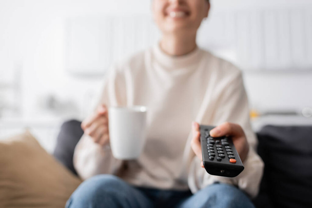 bijgesneden weergave van lachende vrouw met kopje thee klikken tv-kanalen, wazig achtergrond - Foto, afbeelding