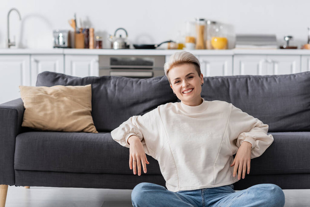 cheerful woman in white pullover sitting near couch in open plan kitchen and looking at camera - 写真・画像