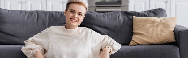 happy woman in white pullover smiling at camera near couch and pillow, banner - Fotografie, Obrázek