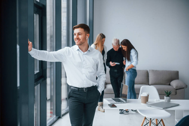 Man in formal clothes standing in front of young successful team that working and communicating together indoors in office. - 写真・画像