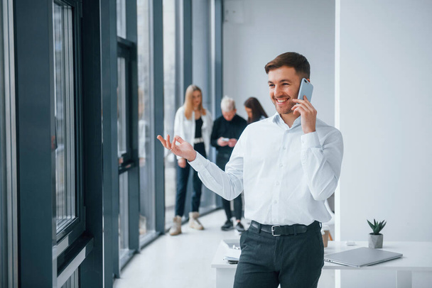 Man in formal clothes talking by phone and standing in front of young successful team that working and communicating together indoors in office. - Фото, зображення