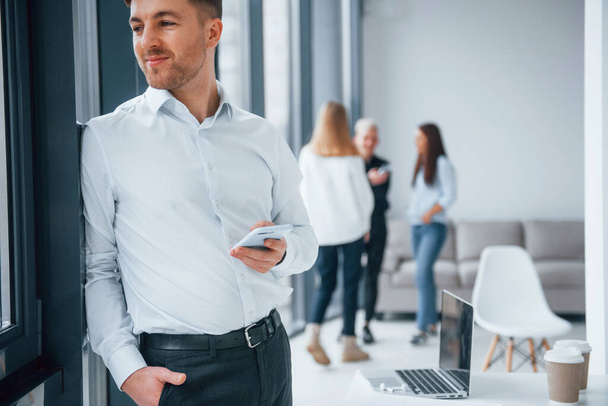 Man in formal clothes standing with phone in hand in front of young successful team that working and communicating together indoors in office. - Foto, Imagem