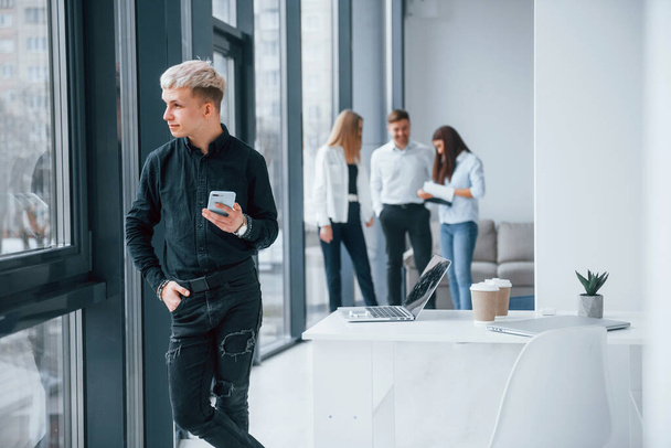 Young hipster with phone standing in front of group of young successful team that working and communicating together indoors in office. - Φωτογραφία, εικόνα