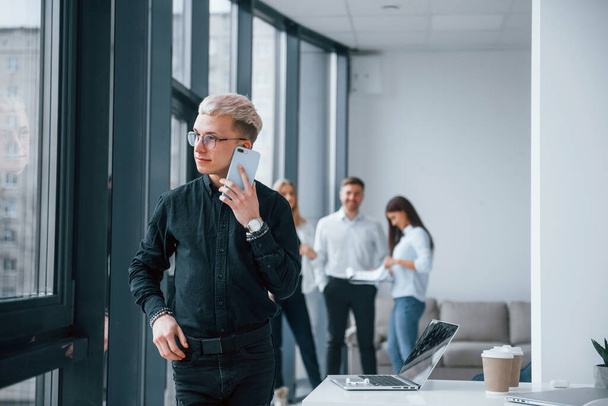 Young hipster with phone standing in front of group of young successful team that working and communicating together indoors in office. - Photo, Image