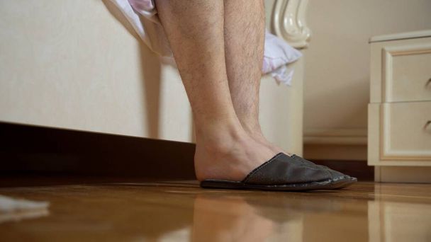 Men's feet shod in slippers near the bed. With one hand, the man holds on to his leg. - Foto, immagini