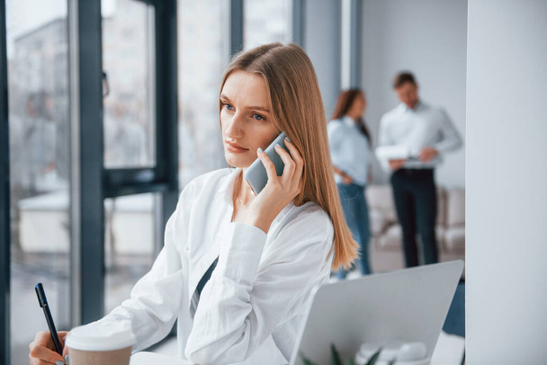 Woman talking by phone in front of group of young successful team that working and communicating together indoors in office. - Photo, Image