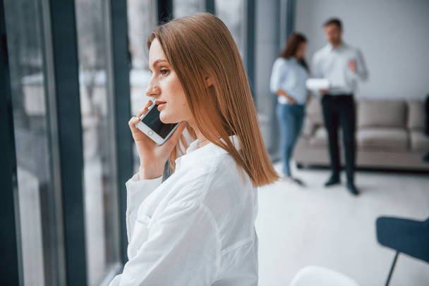 Mujer hablando por teléfono en frente de un grupo de jóvenes equipo exitoso que trabajan y se comunican juntos en el interior de la oficina. - Foto, imagen
