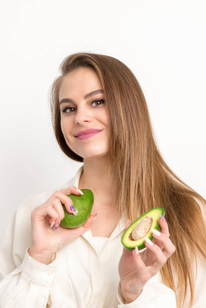 Portrait of a lovely smiling young brunette caucasian woman wearing the white shirt with long hair holding and showing avocado, standing isolated over white background - Zdjęcie, obraz