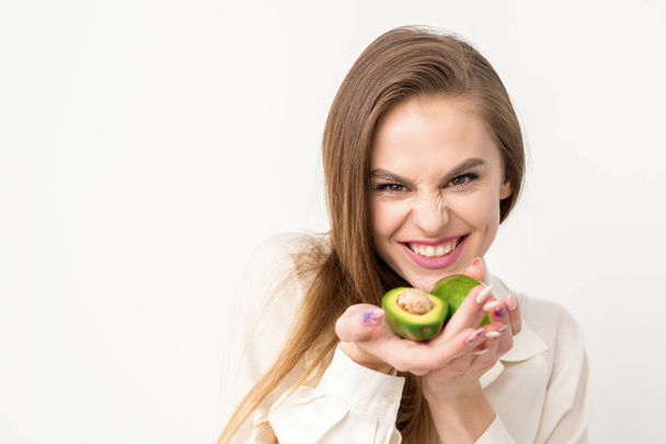 Portrait of a lovely smiling young brunette caucasian woman wearing the white shirt with long hair holding and showing avocado, standing isolated over white background - Photo, Image