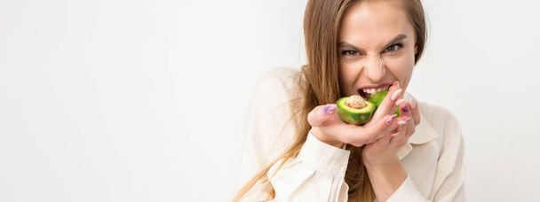 Portrait of a lovely smiling young brunette caucasian woman wearing the white shirt with long hair holding and showing avocado, standing isolated over white background - Photo, Image
