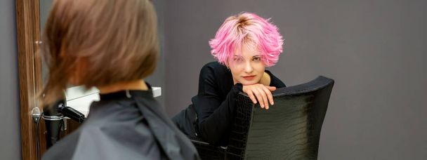 Retrato de una hermosa mujer caucásica joven con un nuevo peinado corto rosa sentado en una silla en un salón de peluquería - Foto, Imagen