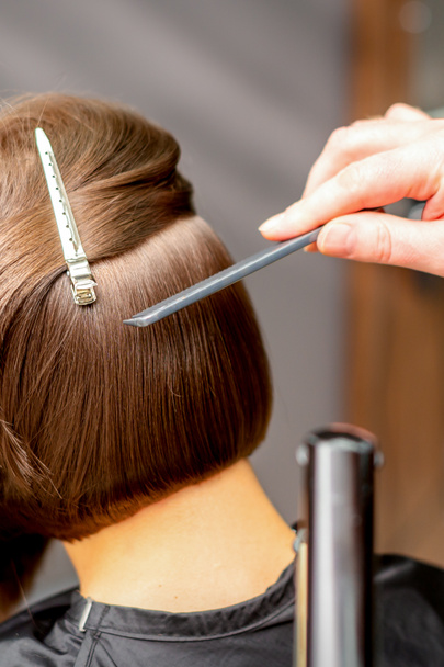 Kadeřnice narovnává krátké vlasy mladé brunetky s plochou žehličkou v kadeřnickém salonu, zblízka - Fotografie, Obrázek