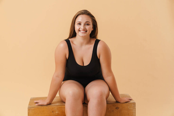 Young woman wearing swimsuit smiling while sitting on wooden cube isolated over beige wall - Zdjęcie, obraz