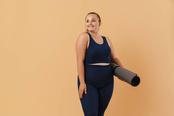 Young woman wearing sport suit smiling while posing with fitness mat isolated over beige background - Фото, зображення