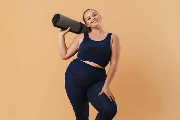 Young woman wearing sport suit smiling while posing with fitness mat isolated over beige background - Φωτογραφία, εικόνα