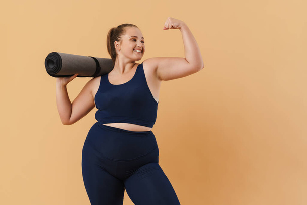 Young woman showing her bicep while posing with fitness mat isolated over beige background - 写真・画像
