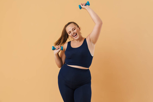 Young woman wearing sport suit doing exercise with dumbbells isolated over beige background - Foto, imagen