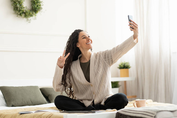 Young smiling woman sitting on bed taking selfie - Foto, immagini