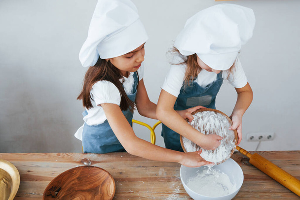 Two little girls in blue chef uniform working with flour on the kitchen. - Fotoğraf, Görsel