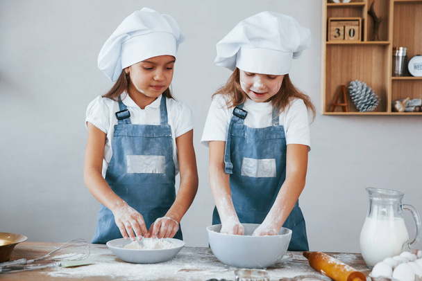 Two little girls in blue chef uniform working with flour on the kitchen. - Фото, изображение