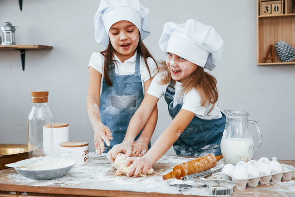 Two little girls in blue chef uniform kneading dough on the kitchen. - 写真・画像