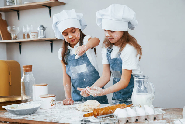 Two little girls in blue chef uniform kneading dough on the kitchen. - Zdjęcie, obraz