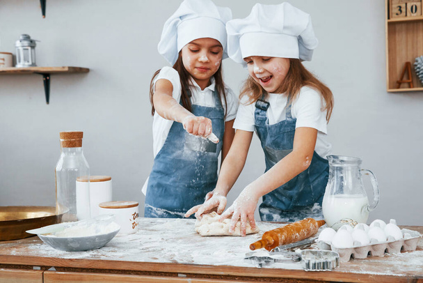 Two little girls in blue chef uniform kneading dough on the kitchen. - Foto, imagen