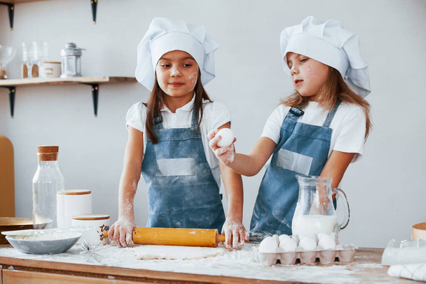 Two little girls in blue chef uniform kneading dough on the kitchen. - Foto, Imagen
